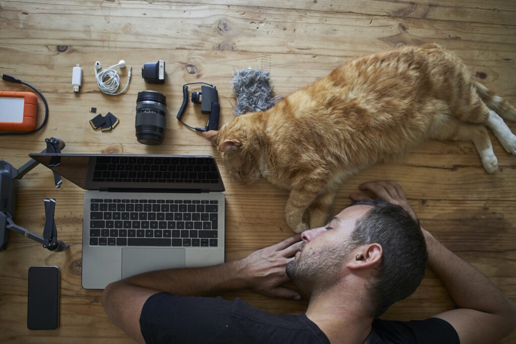 Exhausted man sleeping on table with ginger cat, laptop and photografic equipment